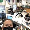 a male teacher with a black face mask takes a selfie of his classroom with remote students on a computer screen, and other students sitting six feet apart with masks at desks