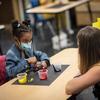 A little girl mixes paint in a classroom wearing a mask. 
