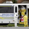 People in hazmat suits load an older man wearing a red robe and a mask into a white transportation bus that says Middlesex County Public Health And Safety