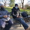 Two men wearing masks sit next to each other on a bench in a park.