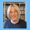 A photo of Bernie Wagenblast, a woman with glasses and blonde bob on a light blue background. In the photo, she sits in front of a bookshelf. She is smiling.