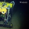 A yellow rover made of wires and wheels sits on the dark seafloor