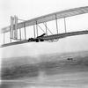 Wilbur takes wing in the 1902 glider soon after the brothers’ return to Kitty Hawk in 1903. Their camp and shed stand alone in the distant wind-swept sands. 