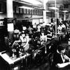 View of a work room at full operation. Men and women work at various stations.