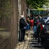 people of color wearing face masks line up outside of a building along a wire fence