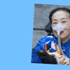 Alice Wong in a power chair. She wears a blue shirt with a pattern of orange, black, white, and yellow lines and cubes. She's masked, but smiling with red lips. The mask is attached to a gray tube.