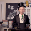 a man in a top hat and wearing a fly tie is at his home doing a remote online awards ceremony. behind him is a poster that reads Ig.