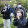 a woman with pepper hair and glasses and wearing a face mask, crouches a little as she looks through binoculars at birds in a park. behind her are a group of fellow birders holding binoculars and wear