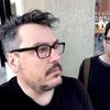 two men both wear black clothing and glasses. John Flansburgh has a goatee and is looks off to the right. John Linnell is staring directly at the camera, looks quite like Alison Bechdel