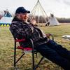 a man sits on a lawn chair in a field with tipis in the background