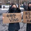 two asian women wearing masks and jackets outside in the snow carrying cardboard signs that say 'hate is a virus' and 'stop asian hate'