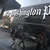 A sign on a wall outside reads 'Washington Post.' Commuters can we be seen in the reflection of the wall.