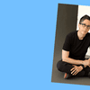 Alison Bechdel sitting cross legged on the floor. She is wearing glasses, a black t-shirt and pants, and has a short haircut. 