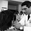 a black and white photo of a male doctor with glasses wearing a white coat with a patch that reads NIAID is examining a patient whose back is to the camera. other clinicians are in the office watching