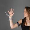 a white woman holds up her right hand. a prosthetic additional thumb is attached to the opposite side of her right hand, accompanied by some wires leading to a strap around her bicep