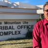 an older indigenous man wearing a red button down shirt and traditional bolo tie and sunglasses stands outside of a building with a sign that reads 'tribal office complex'