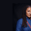 Lynn Nottage, a Black woman with dreadlocks, looks curiously at the camera. She is wearing a denim shirt and a thick, red beaded necklace.