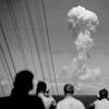 Four men in the foreground watch a tall, which mushroom cloud.