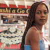 Fey, a Black woman with long dark brown box braids, sits at a counter. She looks at the camera and wears sunglasses on her head and a light blue dress. A black purse is on the counter in front of her.
