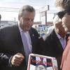 Chris Christie leans slightly over to sign a copy of Time, with the words 'The Boss' visible under his portrait. The magazine is being offered by a man in a pinkish-orange shirt with sunglasses. 