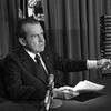 In black and white, Richard Nixon sits at a table in front of a microphone, pointing with his left hand to a pile of binders that can be seen stacked on a table next to his. 