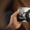 Hrishikesh Hirway’s hand holds a black & white photo of his late mom, Kanta. She’s visible from her neck up in the photo, and in the background of his hand is a wooden table with eyeglasses on top.