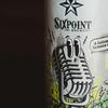 a closeup of a beer can. on the label is a old-school microphone with cartoon eyes looking to the left, it is also whistling. Behind it is a field of plants and a city skyline. the can says 'sixpoint 