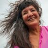 A view of Sandra Cisneros's face and shoulders from below. She is outside, wearing a hot pink blouse, and her long black hair splays behind her in the air while she moves, smiling.