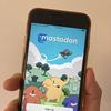 A hand holds an Iphone with the word 'Mastodon' in cartoon letters on a blue sky on the screen. Also on screen is a cartoon plane, with cartoon mastodons on green hills with a login and signup button