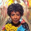 Helga Davis is centered, looking directly toward the camera in front of a bright, colorful tunnel. She wears a garment made of a stunning tangle of twisting, colorful beads.