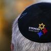 A black kippah on a man's head is shown with the words 'stronger than hate' embroidered in white, next to a golden Star of David and two diamonds, one in blue and one in red. 