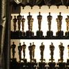 A close-up photograph of a two-tiered shelf displaying dozens of Oscar awards lined up in pairs, framed by a shiny gold curtain and backlit from a display light behind the shelf.