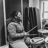 A young woman plays drums while another looks on.