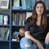 Kate Bowler, a white woman with long brown hair, sits in front of a dark blue bookcase on a blue velvet chair. She wears red lipstick, a navy blue shirt, and light blue jeans.