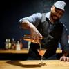 Chris Marshall stands at a wooden bar top and pours a non-alchololic cocktail into a martini glass from a martini shaker.