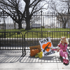 A photograph of a lone protestor outside the White House wearing a pig mask, blonde wig, and pink tracksuit, draped in the American flag, sitting next to a sign reading, War Addicts.