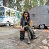 A photo portrait of playwright Larrisa Fasthorse seated on a folding chair seated in a wooded outdoor area with a camper and a bus visible in the background.