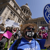 A photo of a pro-abortion protester, wearing a straw hat and dark sunglasses with a facemask reading 'vote,' a shirt reading 'organize' and a sign reading 'keep abortion legal' with other protesters. 
