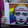 A photo of what looks like a protester, holding up a sign depicting Russian president Vladimir Putin in drag makeup with a pride flag behind him along with the words, stop homophobia.