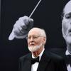 A photographed portrait of film composer, John Williams, wearing a black tuxedo, standing in front of a large printed portrait of himself when he was younger, also wearing a black tuxedo.