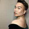 A photographed portrait of poet Paul Tran, wearing a dark velvet top, winged eye makeup, a short haircut, and a septum piercing, posed looking toward the camera over their shoulder.
