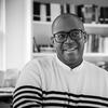 A black and white photo of writer Brandon Taylor, a Black man wearing thick rimmed glasses and a striped sweater, sitting near a bookshelf.