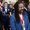 A photographed portrait of writer Colson Whitehead from an award ceremony, wearing long dreadlocks, light brown glasses, a light blue suit, and a medal with a red ribbon, and a crowd behind him.