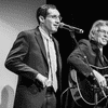 A singer, standing, and a seated guitarist performing onstage