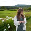 A photograph of Kate Beaton, a white, brunette with long hair, standing in front of a rolling green meadow on a summer day. She’s wearing a black and white dress and a white cardigan.