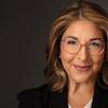 A photographed portrait of writer Naomi Klein, wearing transparent glasses, gold hoop earrings, and a black blazer. 