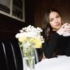 A photo portrait of singer Olivia Rodrigo, wearing a black sweater, holding a white mug, sitting at a table with a small arrangement of flowers in the foreground.