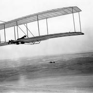 Wilbur takes wing in the 1902 glider soon after the brothers’ return to Kitty Hawk in 1903. Their camp and shed stand alone in the distant wind-swept sands.