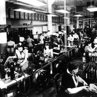 View of a work room at full operation. Men and women work at various stations.
