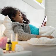 Sick girl with flu lying in bed at home, checking temperature with thermometer and on her tablet